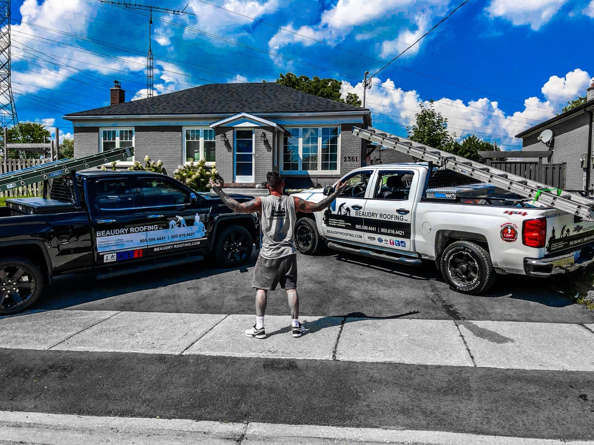 Justin with arms spread between two Beaudry Roofing trucks in front of a bungalow