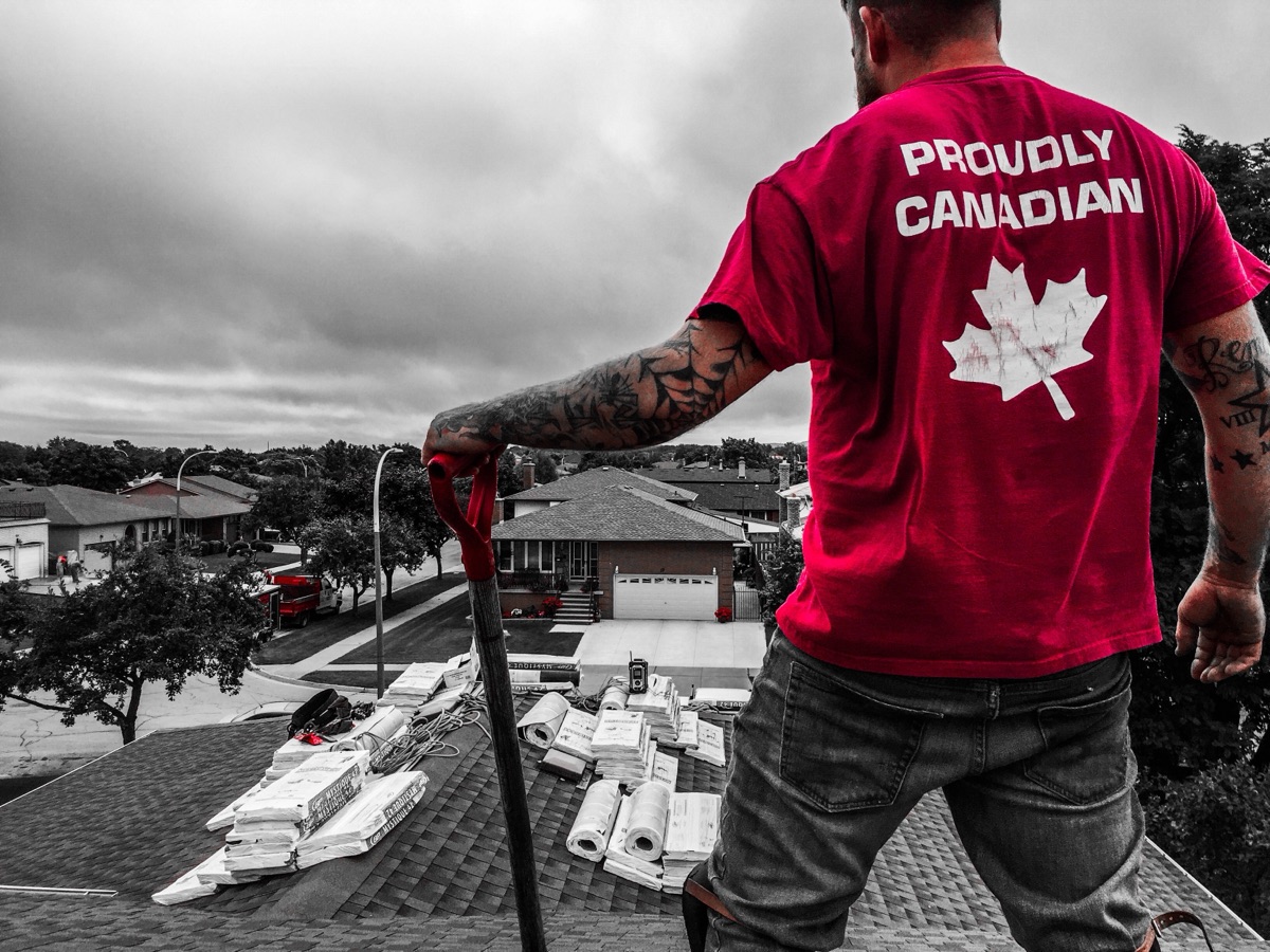 Justin on a roof in a Proudly Canadian shirt, shingle bundles staged along the ridge