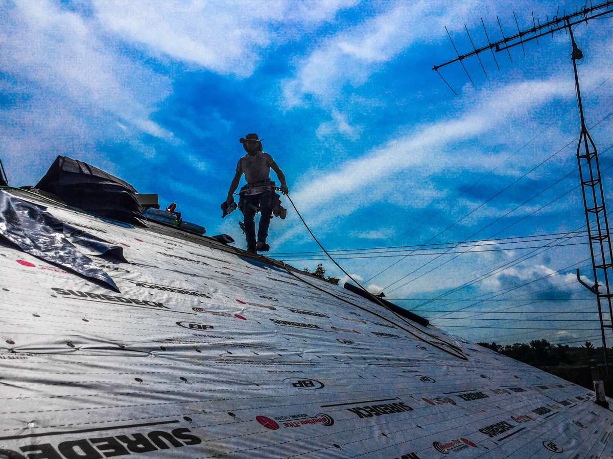 Roofer standing on the peak of a roof wrapped in synthetic underlayment against a blue sky