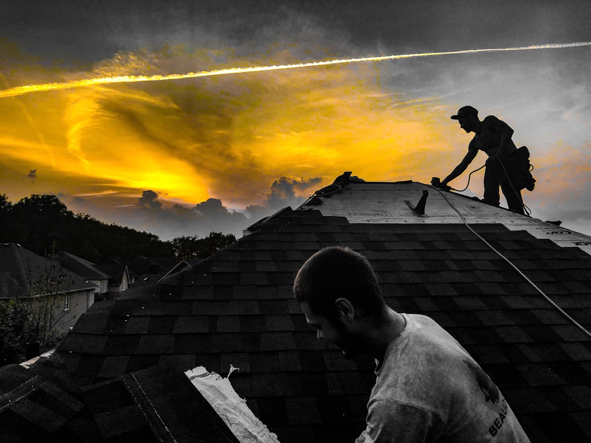 Two Beaudry roofers laying shingle courses on a hip roof under a sunset sky